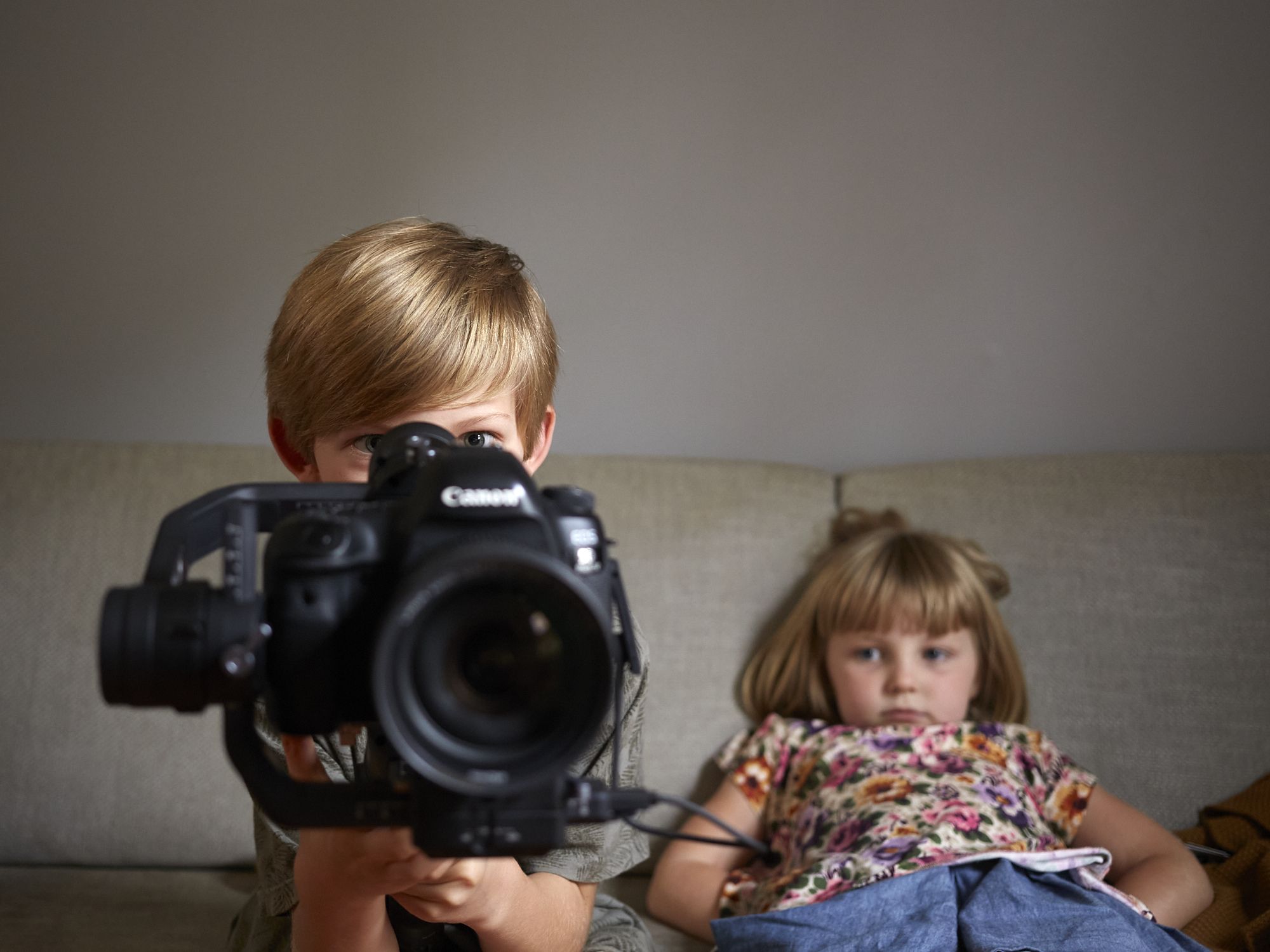 Two kids of a couch, sitting behind a big camera.